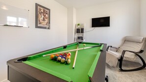Ett biljardbord med bollar, en whiteboard, en inramad tavla, en tv och en fåtölj i ett rum med vita väggar.