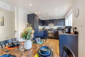 A modern kitchen with dark blue cabinets, a wooden dining table set with plates and glasses, and a vase with flowers. - 3 Bed in Aspatria (oc-h38206) (Aspatria)