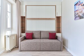Een sofa in neutrale tinten, met twee rode kussens, een radiator en een raam met gordijnen.