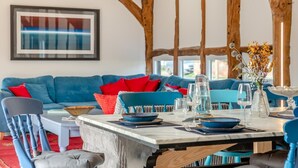 A dining area with a table set for four, a blue sofa in the background, and wooden beams on the ceiling. - 3 Bed in Eastbury (oc-t38204) (Eastbury)