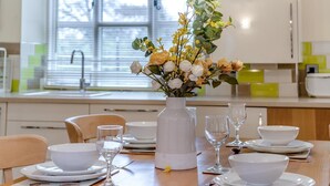 Une table à manger dressée avec de l’assiette blanche, des bols et des verres à vin, un vase blanc rempli de fleurs et, en arrière-plan, une fenêtre avec des stores.