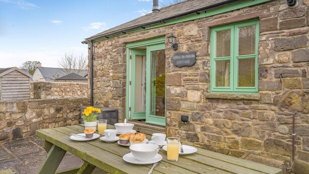 Un bâtiment en pierre avec une porte verte et des volets aux fenêtres, une table de pique-nique en bois avec des aliments pour le petit-déjeuner et un mur en pierre.