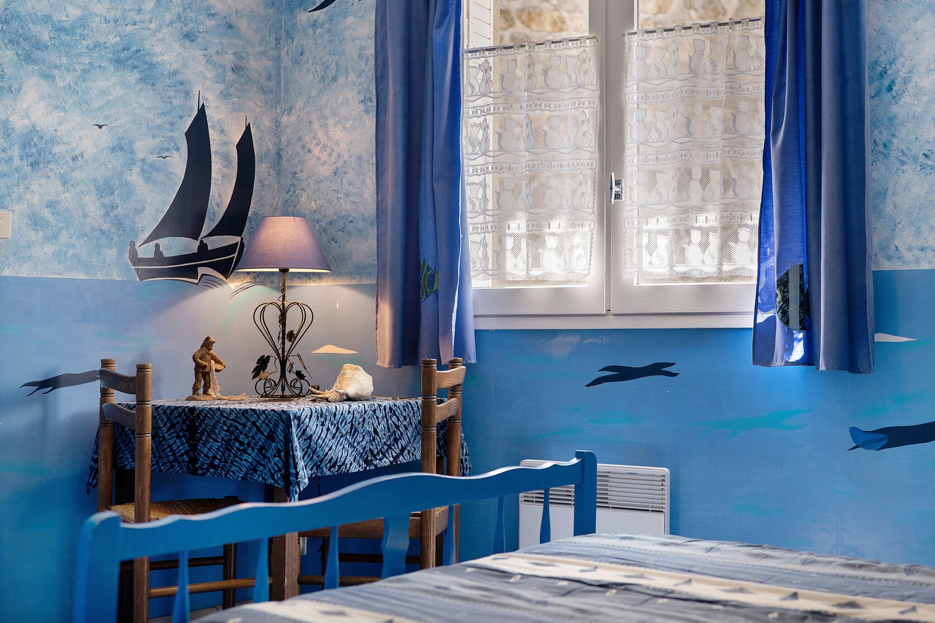 A room with a table, chairs, a lamp, a window with curtains, and wall decorations including a sailboat and fish.