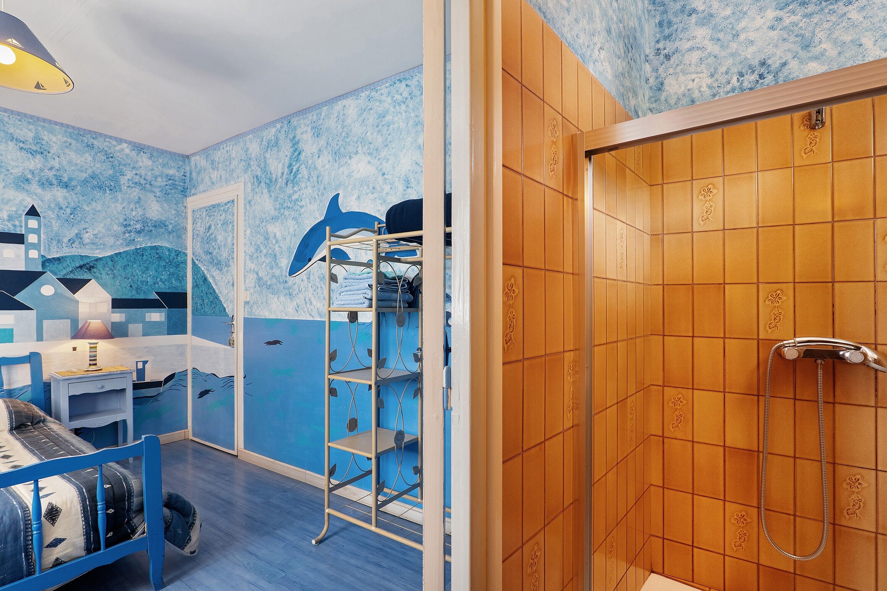 Une chambre avec un mur décoré dans le style marin, aux couleurs bleu et blanc, une porte en bois donnant sur une douche, et un lit avec une tête de lit bleue.