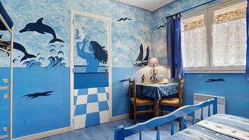 Une pièce décorée sur le thème de la marine, avec des fresques murales représentant des dauphins et des voiliers, un sol à carreaux, une table avec une lampe et une fenêtre avec des rideaux.