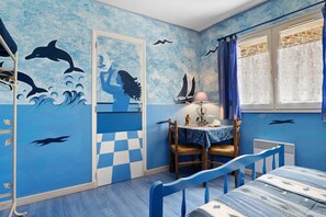 Een kamer met een nautisch thema, met muurschilderingen van dolfijnen en zeilboten, een geruit vloertje, een tafel met een lamp en een raam met gordijnen.