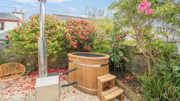 Un bain à remous en bois, doté d’une structure métallique, entouré d’un jardin planté de fleurs et bordé d’un mur en pierre.