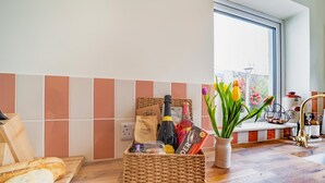 Une cuisine avec un mur recouvert de carreaux, un panier en osier contenant des aliments, une fenêtre ornée de fleurs et une planche à découper en bois avec du pain.