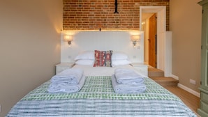 Une chambre d’hôtel avec un mur en briques, une tête de lit blanche, deux tables de chevet avec des lampes, un lit recouvert d’une couvre-lit à motifs et une porte sur la droite.
