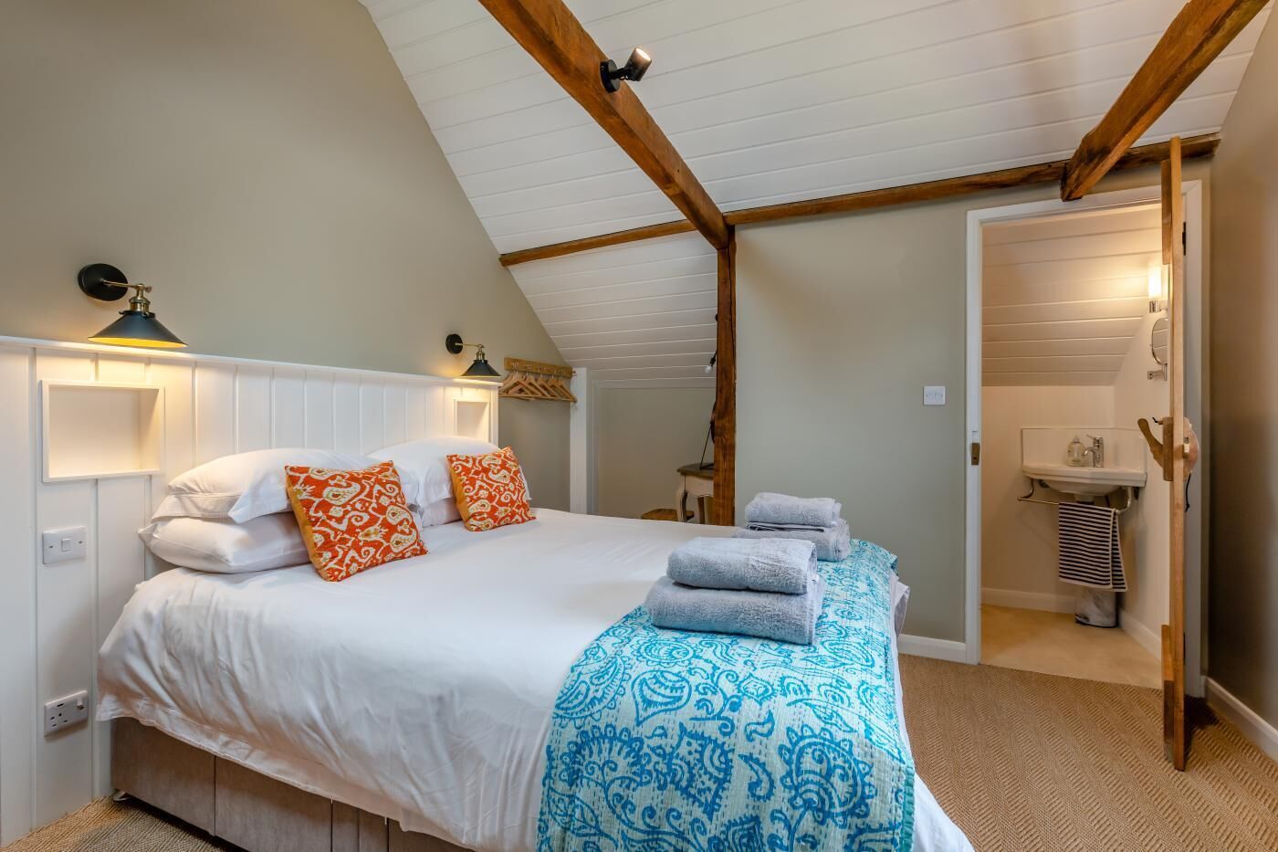 Quarto com cama, almofadas, luminária de cabeceira, porta-toalhas e uma porta de acesso a um banheiro.