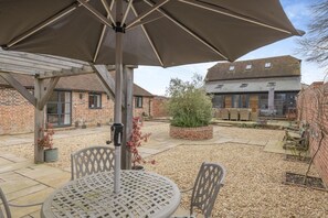 A patio area with a table and chairs, a large umbrella, and a brick building in the background. - 3 Bed in Peasmarsh (oc-graohf) (Peasmarsh)