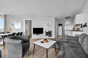 A modern living room with a flat-screen TV, grey sofas, a small white table with fruit and remote controls, and a fireplace. - MeerWeite Holiday Apartment: Luxury & Stunning Sea Views Right by the Coast (Wendtorf)