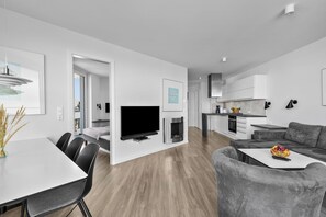 Ein moderner Wohnraum mit Essbereich, Flachbildfernseher, Kamin und einer Küche mit Einbauofen, sowie einem grauen Sofa.