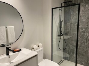 Un baño moderno con espejo redondo, lavamanos blanco, inodoro y ducha con mampara de vidrio.