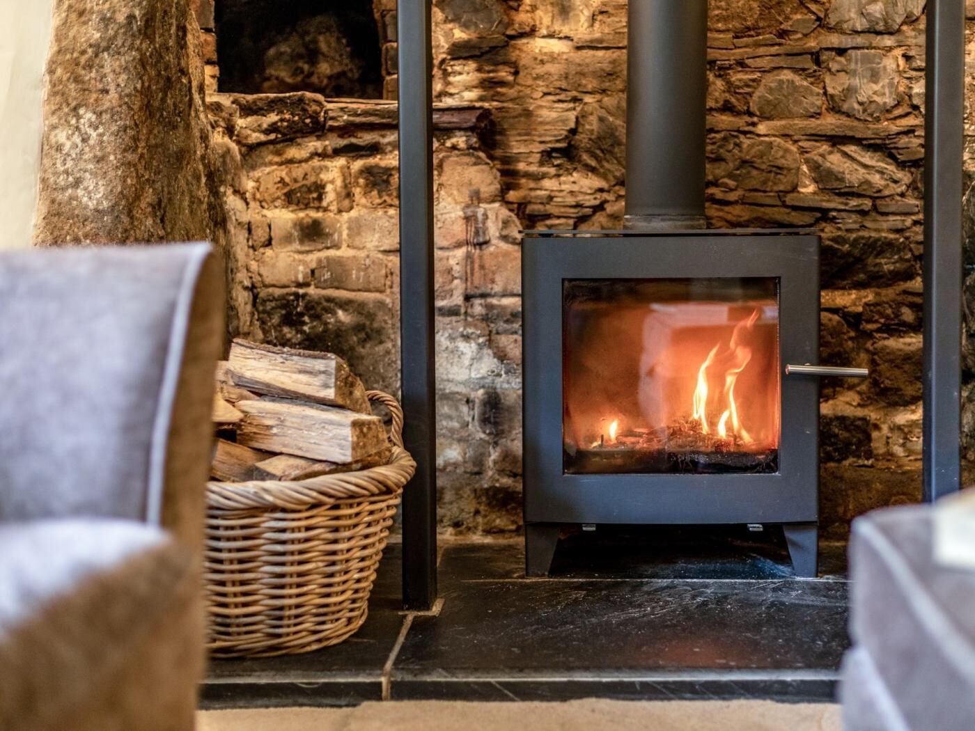 A stone fireplace with a modern wood-burning stove, a wicker basket with logs, and a chair in the foreground.