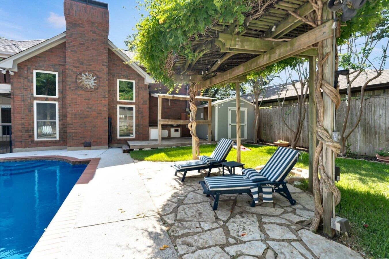 Un patio trasero con piscina, sillas de descanso, una pérgola y una casa de ladrillo.