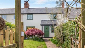 A quaint cottage with a stone pathway leading to a green door, surrounded by lush greenery and a wooden fence. - 3 Bed in Holcombe Rogus (oc-a39001) (Holcombe Rogus)