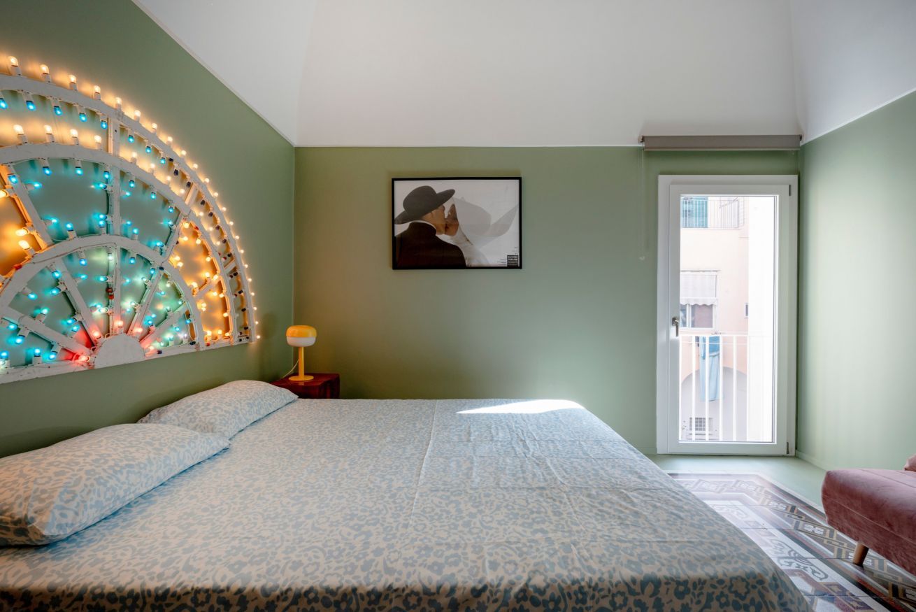 A bedroom with a bed, a wall-mounted light fixture, a framed picture, and a door leading to an outdoor area.