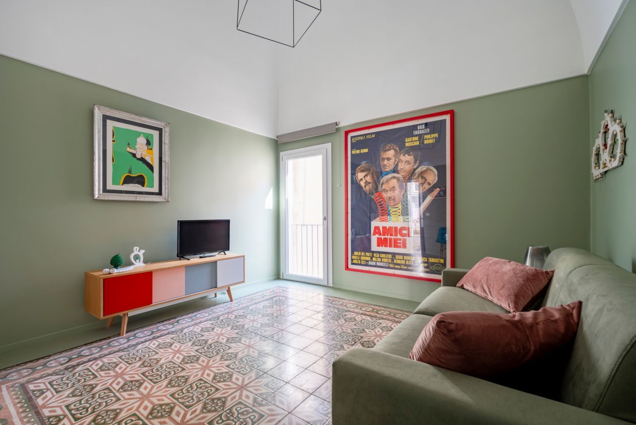 Uma sala de estar com um sofá, uma televisão em um suporte, um piso estampado e um cartaz na parede.