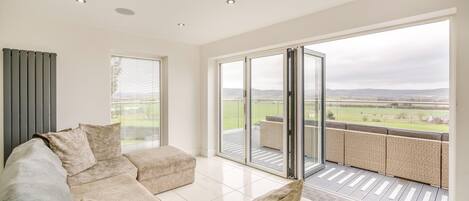 A modern living room with a sectional sofa, a sliding glass door leading to a balcony with a view of the countryside.