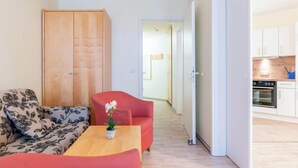 A compact living space with a sofa, a small table with a plant, a wardrobe, and a kitchen area visible through an open door. - Ferienwohnung mit Ahoi-rabatt in Sellin (Sellin)