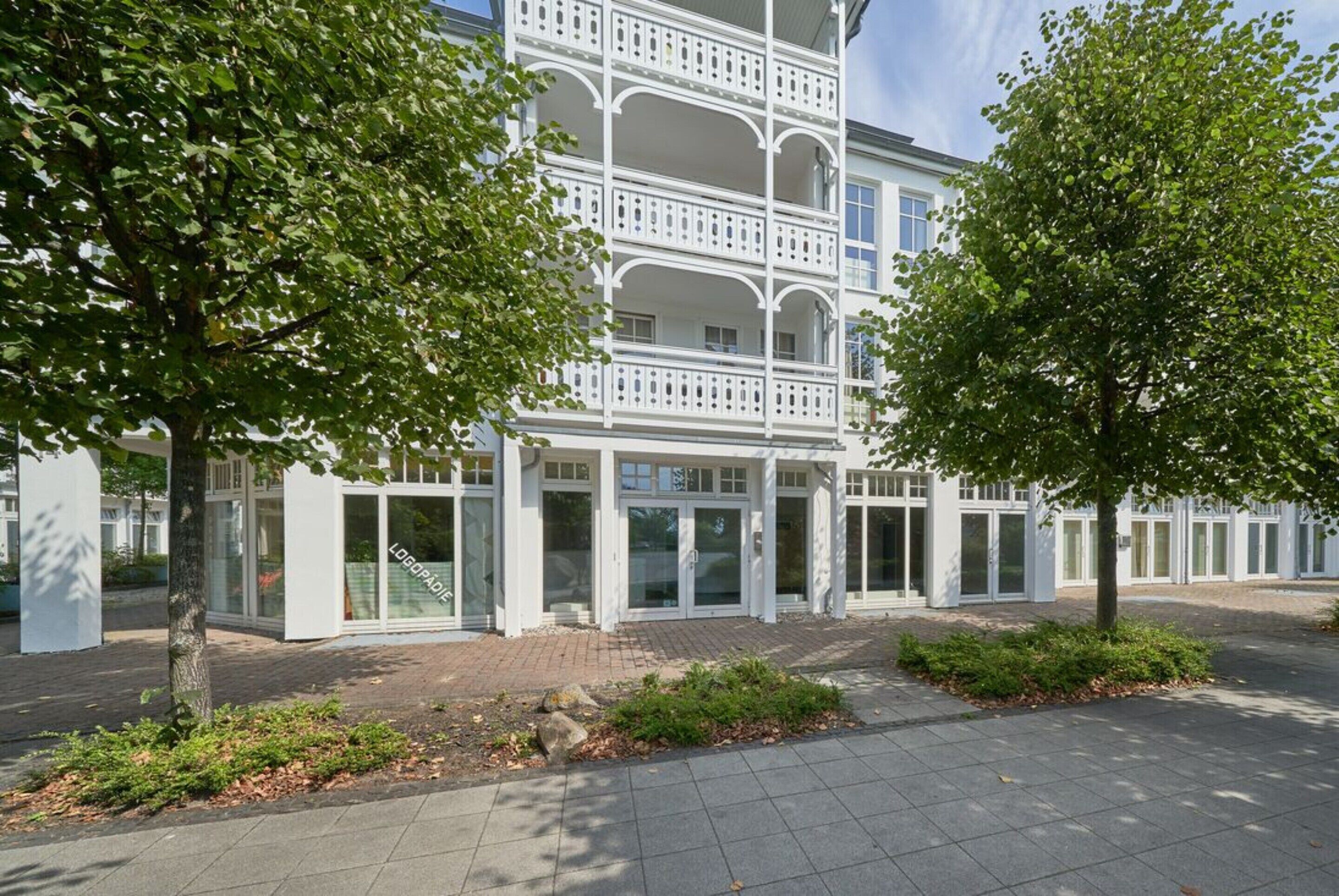 A modern building with a white facade, multiple balconies, and a paved entrance area with greenery.