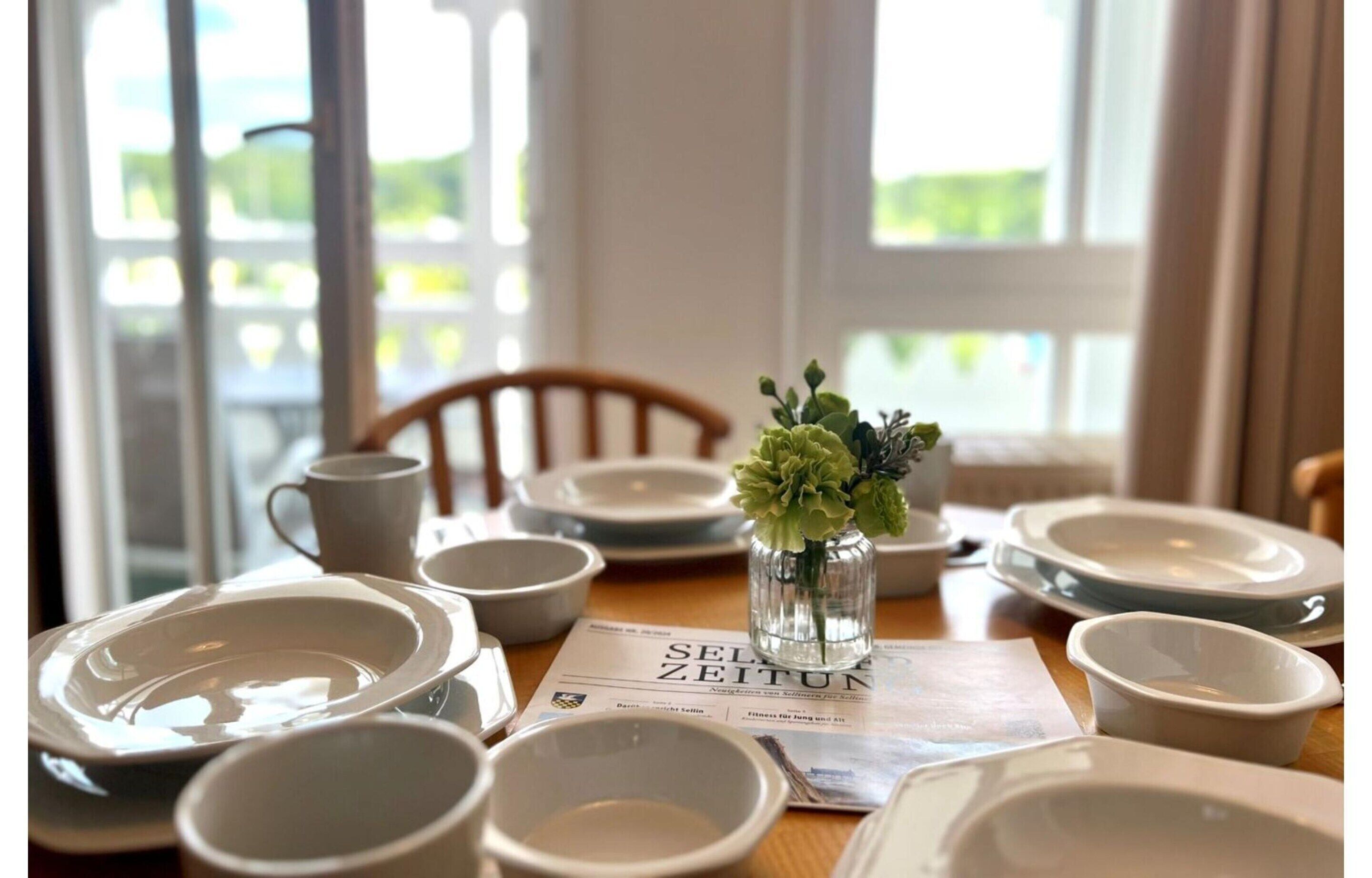 A dining table set with white plates, bowls, and a glass vase with green foliage.