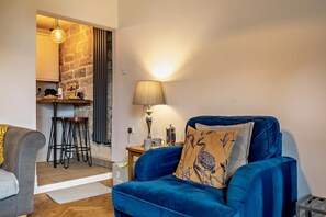 A living room with a blue sofa, a lamp, and a kitchen area with a stone wall. - 1 Bed in Crich (oc-h37533) (Crich)
