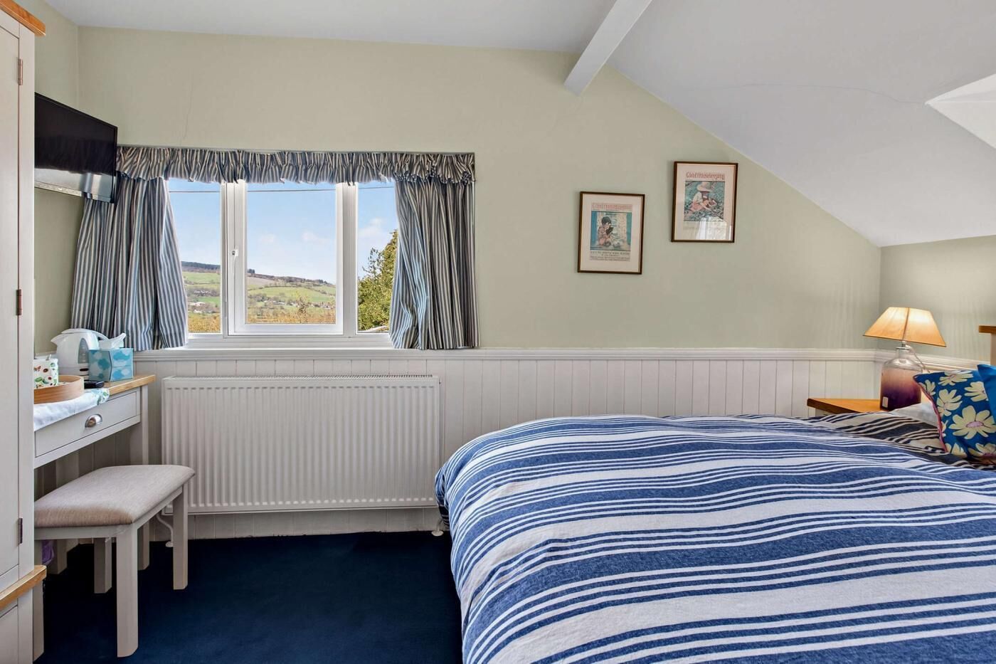 A bedroom with a bed, a desk, a chair, a radiator, a window with curtains, and two framed pictures on the wall.