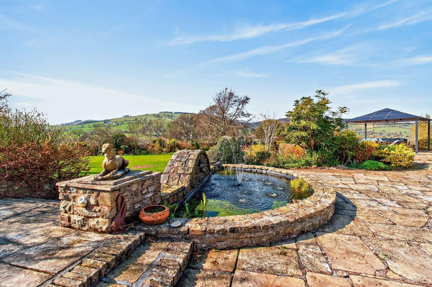Une fontaine en pierre avec un bassin circulaire, un banc en pierre orné d’une sculpture et une pergola dans un jardin aménagé.