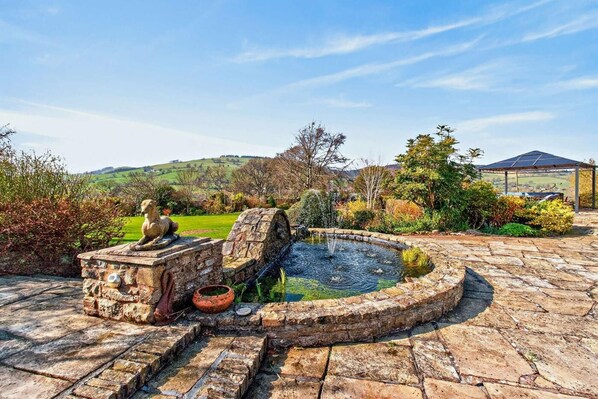 A stone fountain with a circular basin, a stone bench with a sculpture, and a pergola in a landscaped garden.