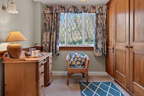 A room with a wooden desk, a chair, a lamp, a window with patterned curtains, and a wooden wardrobe.