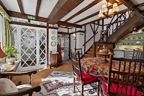 Habitación tradicional con vigas de madera a la vista, una escalera, una zona de comedor con mantel floral y una cocina con estantes llenos de vajilla.
