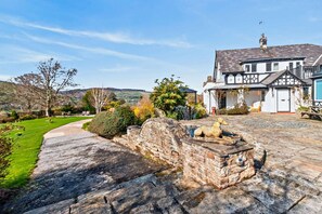 A traditional house with a stone pathway leading to a stone fountain, surrounded by a well-maintained garden and a view of rolling hills. - 5 Bed in Maeshafn (oc-p37882) (Llanferres)