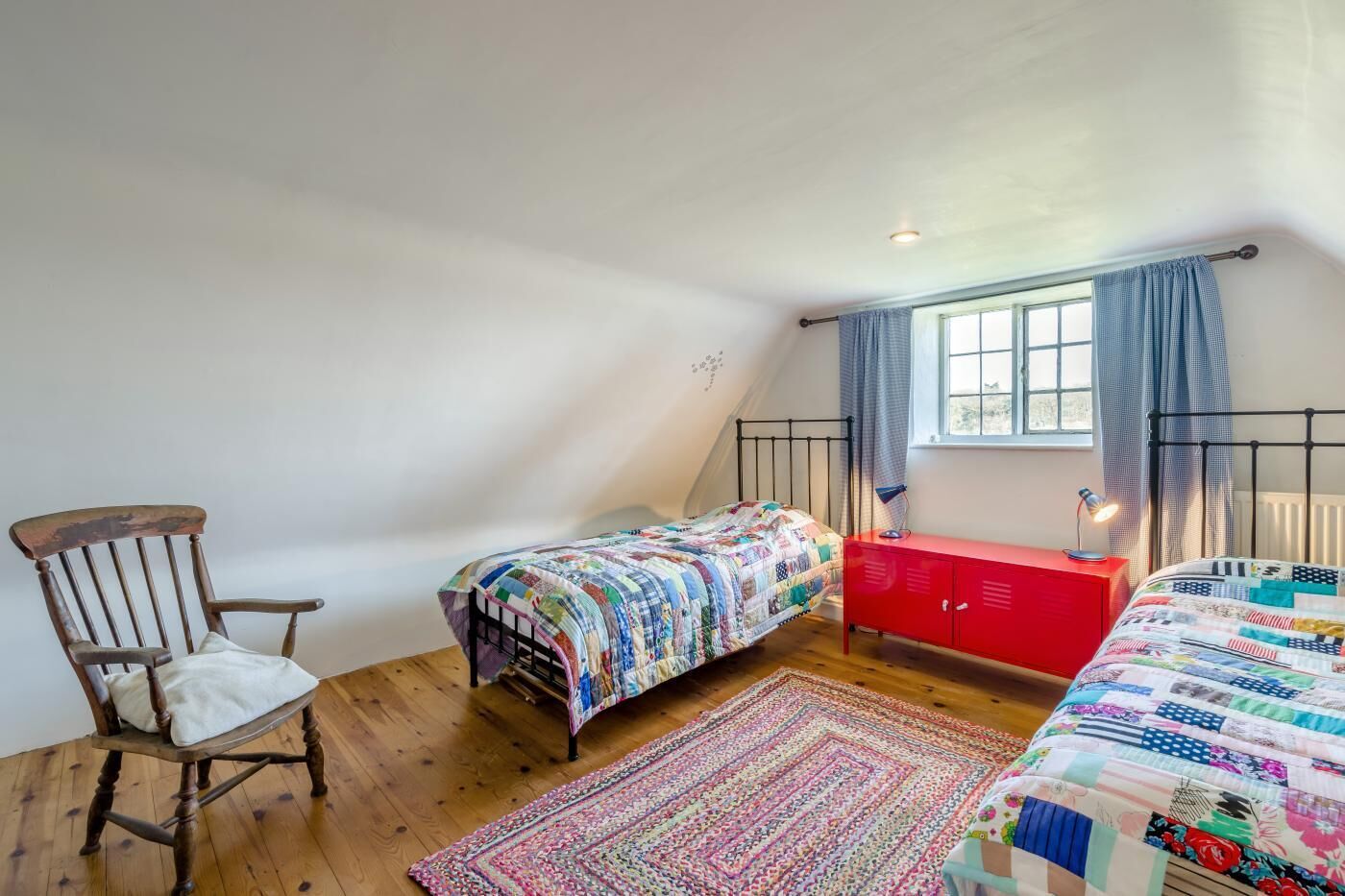 A bedroom with two beds, a red dresser, a wooden chair, and a window with curtains.