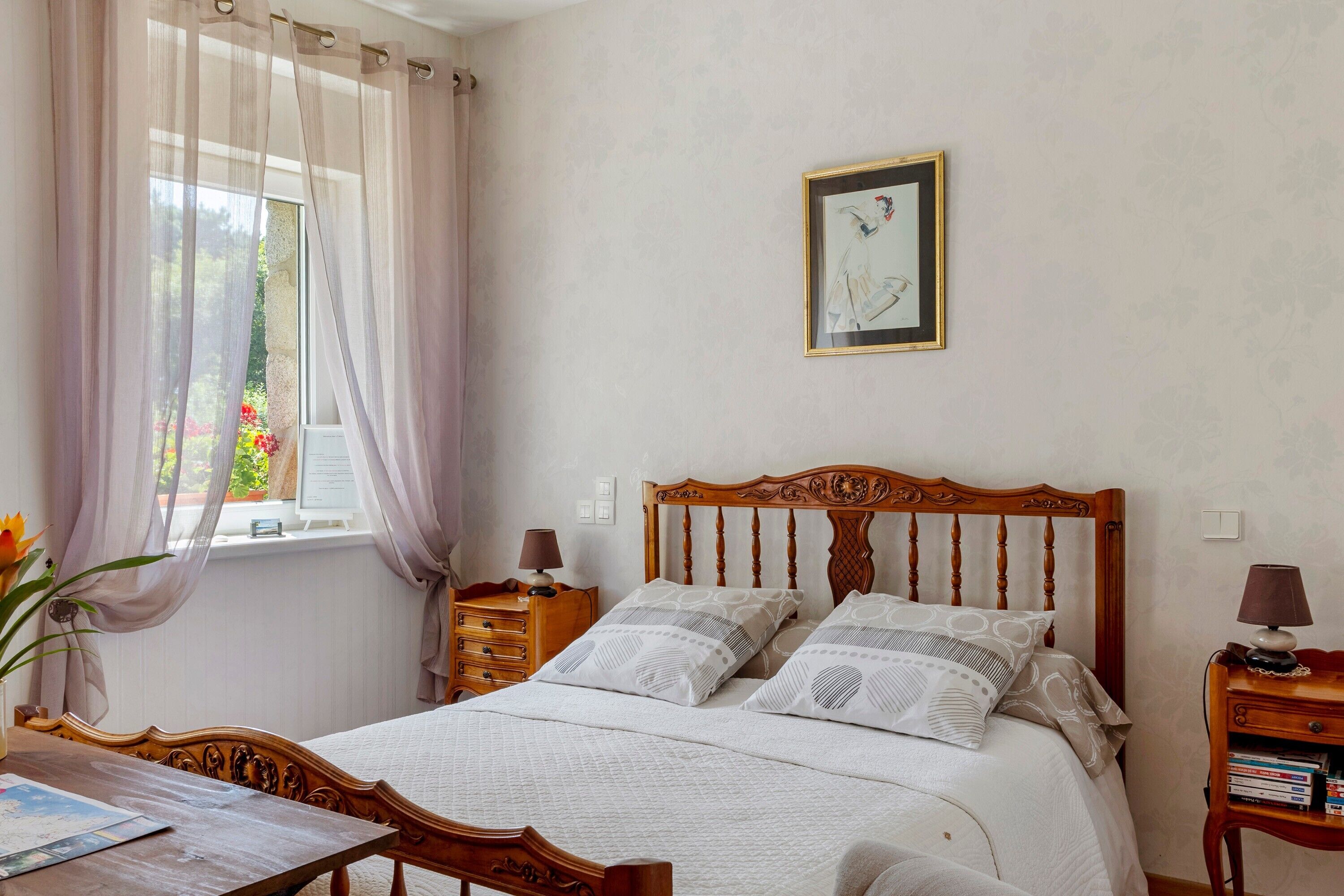 Makuuhuoneessa on puinen sänky, yöpöytä, jossa on lamppu, ikkuna, jossa on läpikuultavat verhot, ja seinällä on kehystetty taulu.