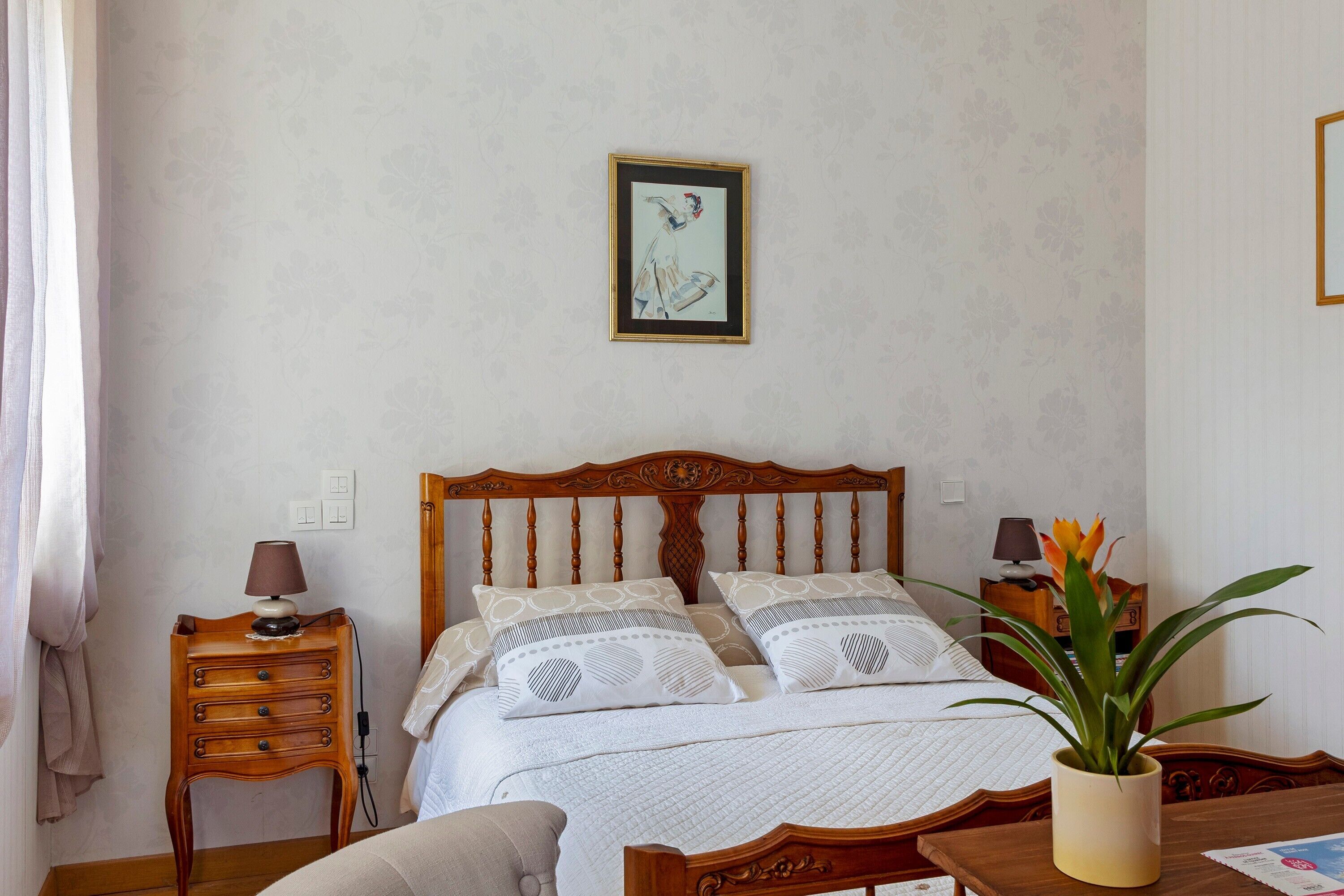 Een slaapkamer met een houten bed, nachtkastjes, een lamp, een potplant en een lijst aan de muur.