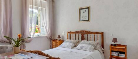 A neatly arranged bedroom with a bed, bedside tables, a window with curtains, a wall-mounted picture, and a small table with a plant.