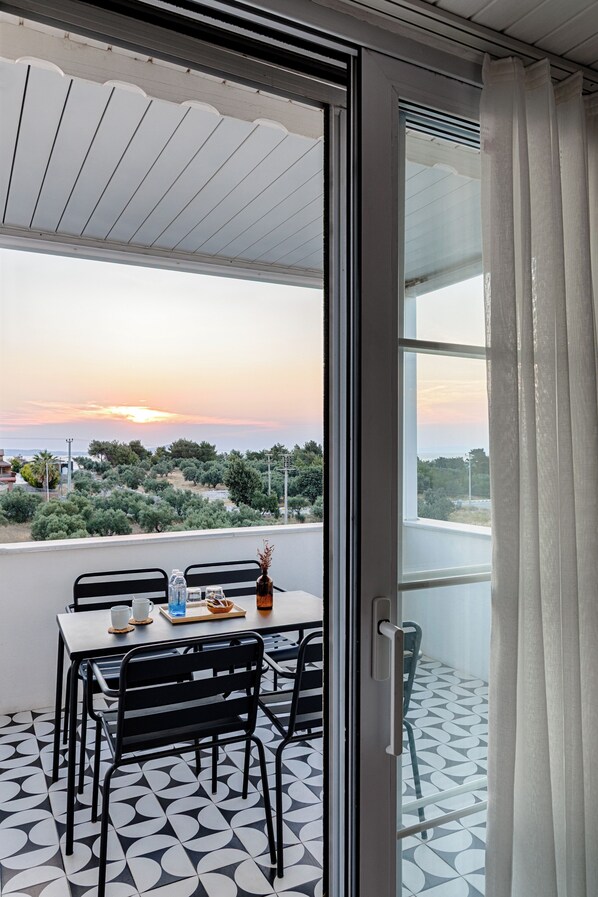 A balcony with a table and chairs, overlooking a landscape with trees and a sunset.