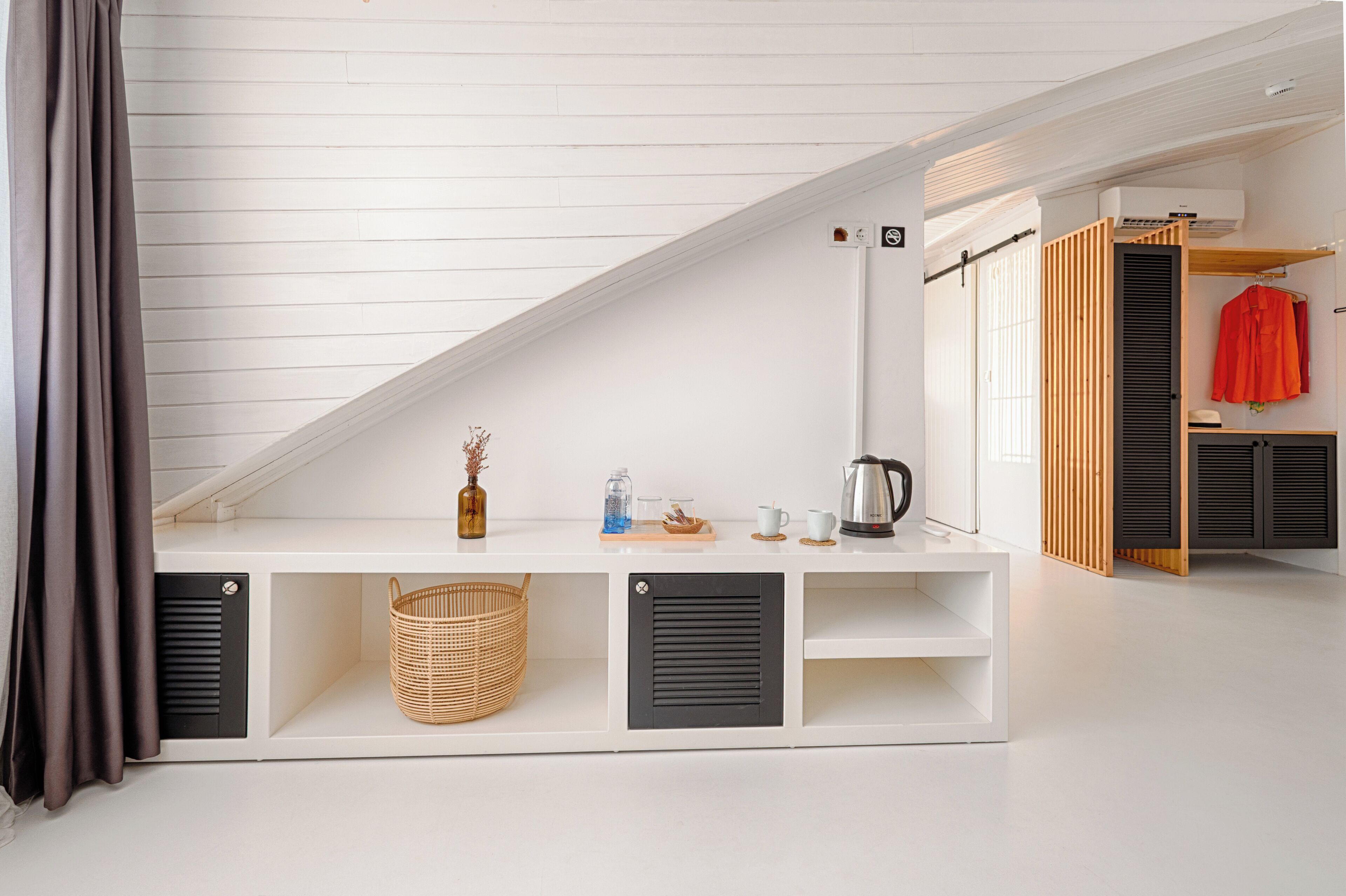 Una habitación con techo inclinado, una mesa consola blanca con espacio de almacenamiento, una cesta de mimbre, un hervidor y una chaqueta roja colgada en un perchero.