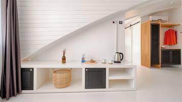 Una habitación con techo inclinado, una mesa consola blanca con espacio de almacenamiento, una cesta de mimbre, un hervidor y una chaqueta roja colgada en un perchero.