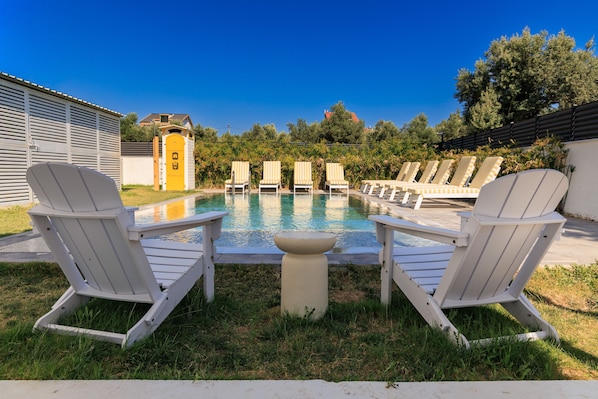 A poolside area with white Adirondack chairs, a small table, and a yellow door in the background.