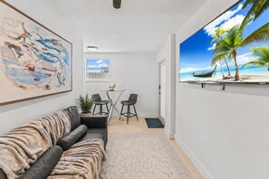 Ein modernes Wohnzimmer mit einer Couch, einem kleinen Tisch mit Stühlen, einem Gemälde und einem großen Fernseher, der eine Strandansicht zeigt.