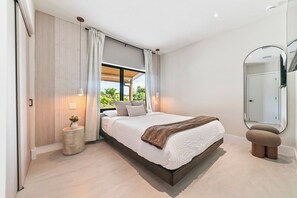 Ein modernes Schlafzimmer mit einem großen Bett, einem kleinen runden Tisch, einem Spiegel und Blick auf Grünfläche.