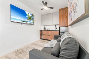 Ein Wohnzimmer mit grauem Sofa, Deckenventilator, Flachbildfernseher, Küchenzeile mit Holzausstattung und einem großen Gemälde an der Wand.