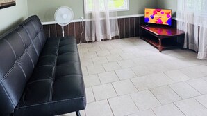 A living room with a black leather sofa, a TV on a wooden stand, a fan, and a window with sheer curtains.