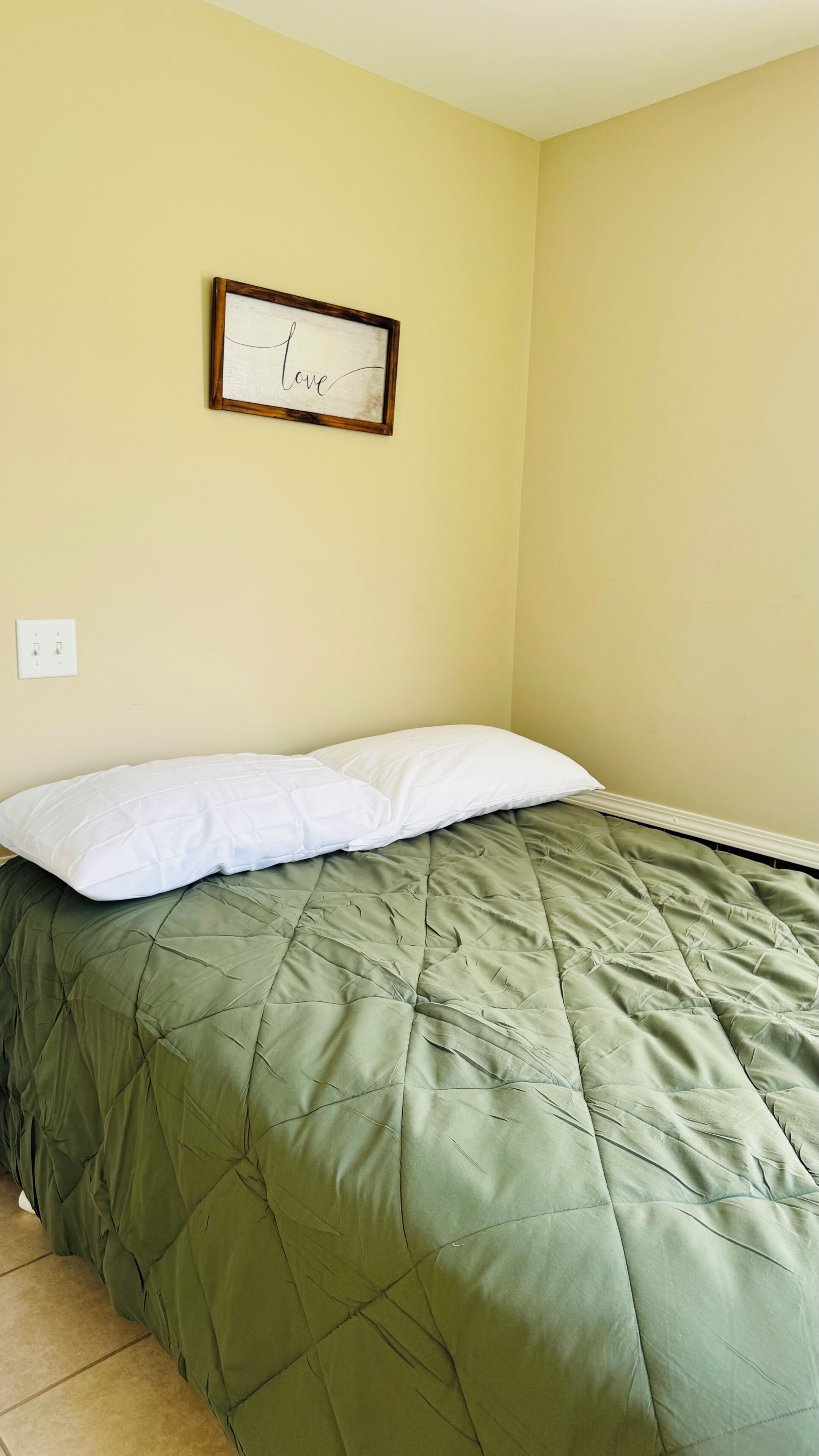 A room with a bed, a wall-mounted light switch, and a framed sign with the word 'love'.