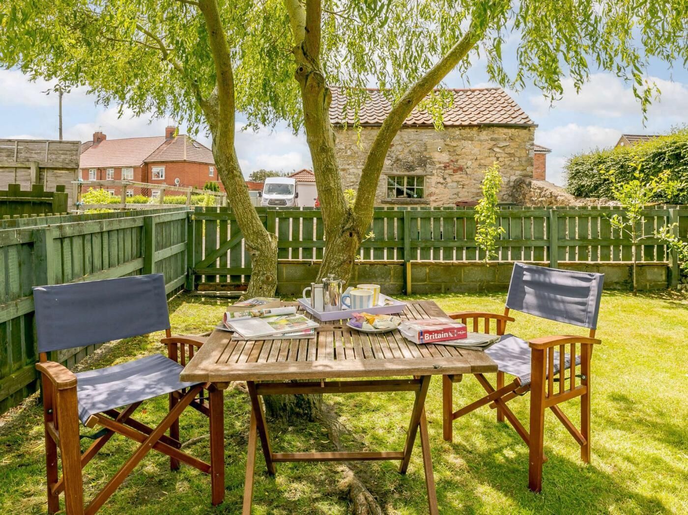A wooden table with chairs in a backyard, surrounded by trees and a stone building.