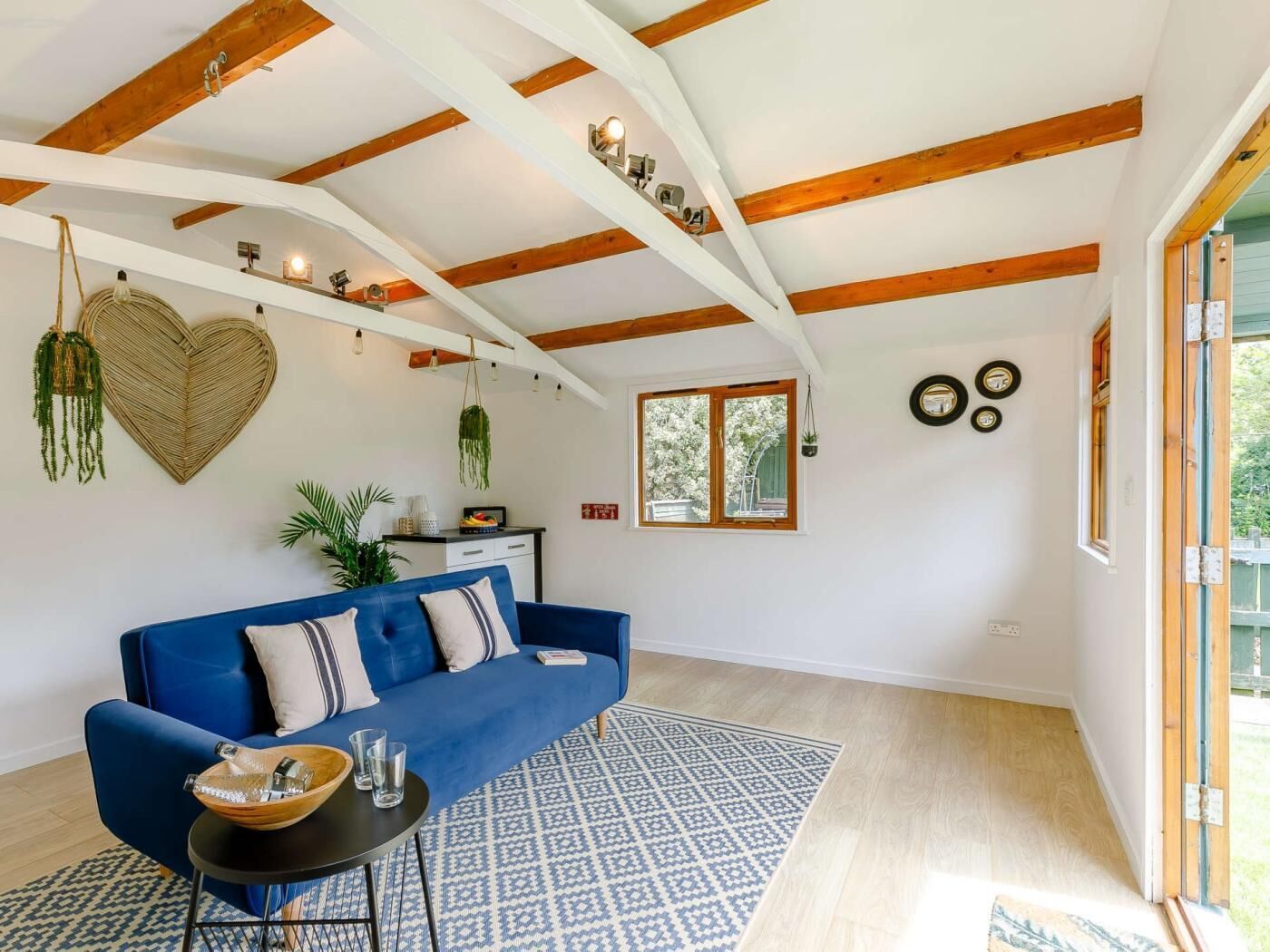 Een moderne woonkamer met een blauwe bank, houten balken, een kamerbreed tapijt met dessin en een hartvormige wanddecoratie.