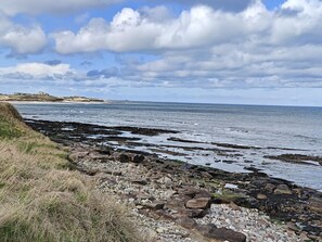 A coastal landscape with rocky shores, a grassy area, and a distant building on a hill. - 2 Bed in Seahouses (oc-p39046) (Seahouses)